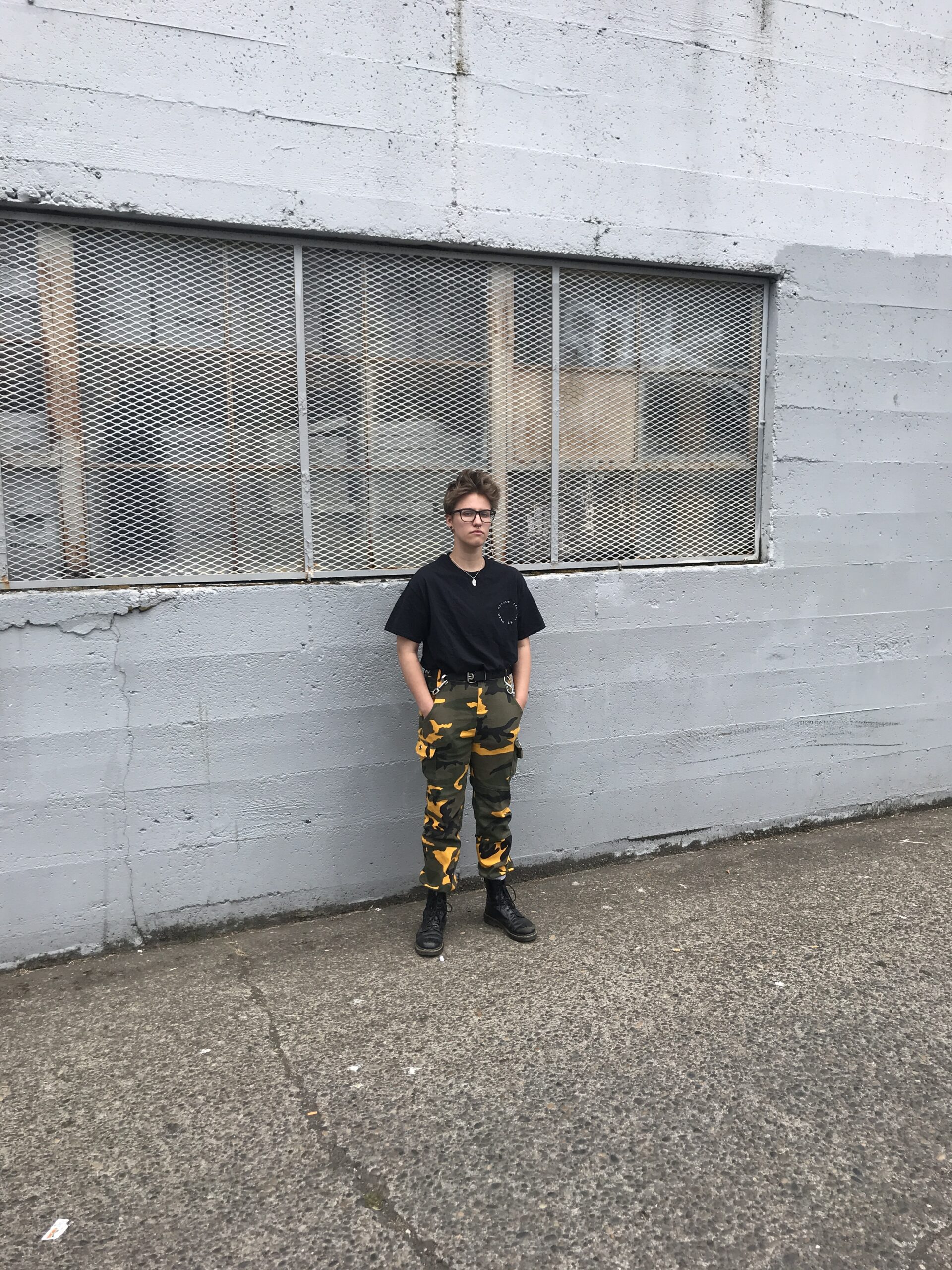 Img of buck, a nonbinary person in camo pants and a black shirt in front of an industrial building.